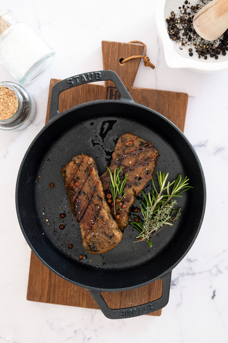 Veganes Wheaty Seitan-Steak mit Röststreifen in einer gusseisernen Pfanne, dekoriert mit Rosmarin, Pfeffer und Salz auf hellem Hintergrund.